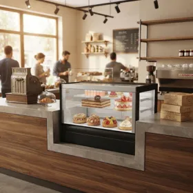 Square glass countertop cake display refrigerator in a bakery shop, presenting layered cakes and pastries in an organized layout.